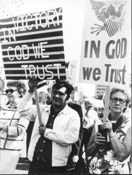 Two demonstrators (a man & a woman) carry signs with the "In God we Trust" theme