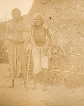 Two unidentified people standing outside grass hut in a village in India